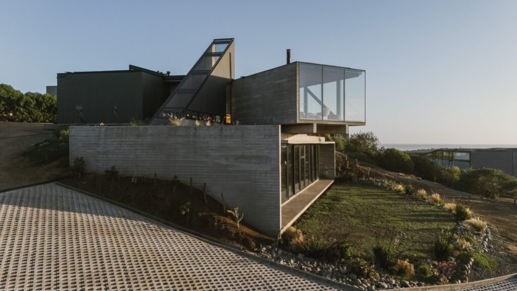 Exterior view of Casa 22 showing rotated upper concrete and glass volumes, a diagonal skylight shaft, and a permeable paved driveway.