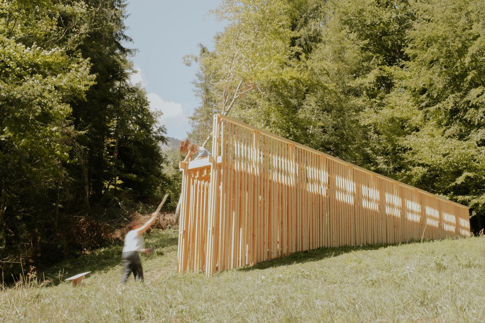 Visitors interacting with a long timber architectural installation along a grassy slope surrounded by lush green forest trees.