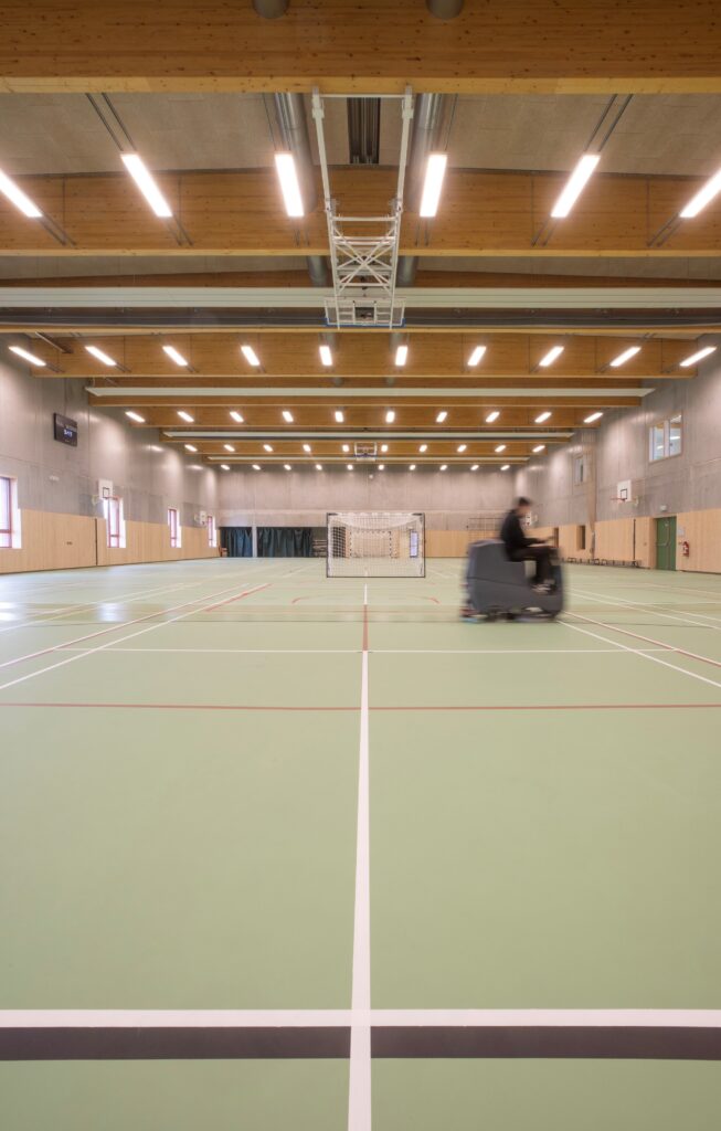 Spacious indoor sports hall with wooden ceiling beams, marked court lines, and high-level lighting.