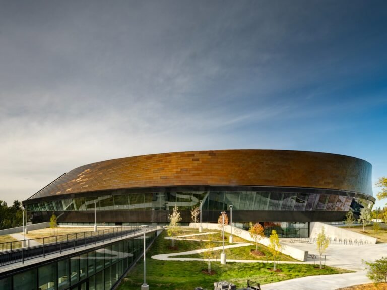 Exterior daytime view of Coronation Park Sports Centre showing the bronze-toned curved facade and the glass link connecting to the Peter Hemingway Aquatic Centre.