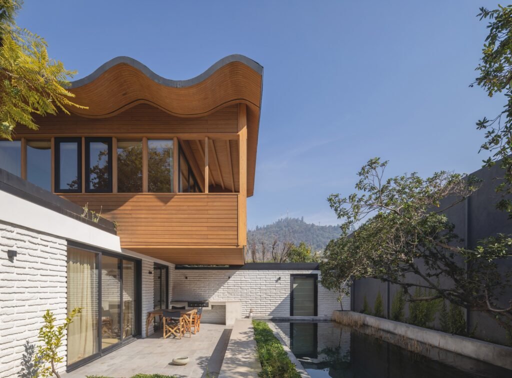 Exterior view of Los Navegantes House showing the upper-floor timber extension with a undulating wavy roof above white brick walls and a natural swimming pool.