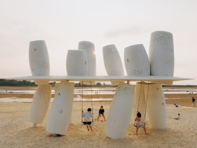 Frontal eye-level view of the Coral Installation featuring perforated white stainless steel cylinders pierced by a thin horizontal roof with children playing on swings on the sandy beach.