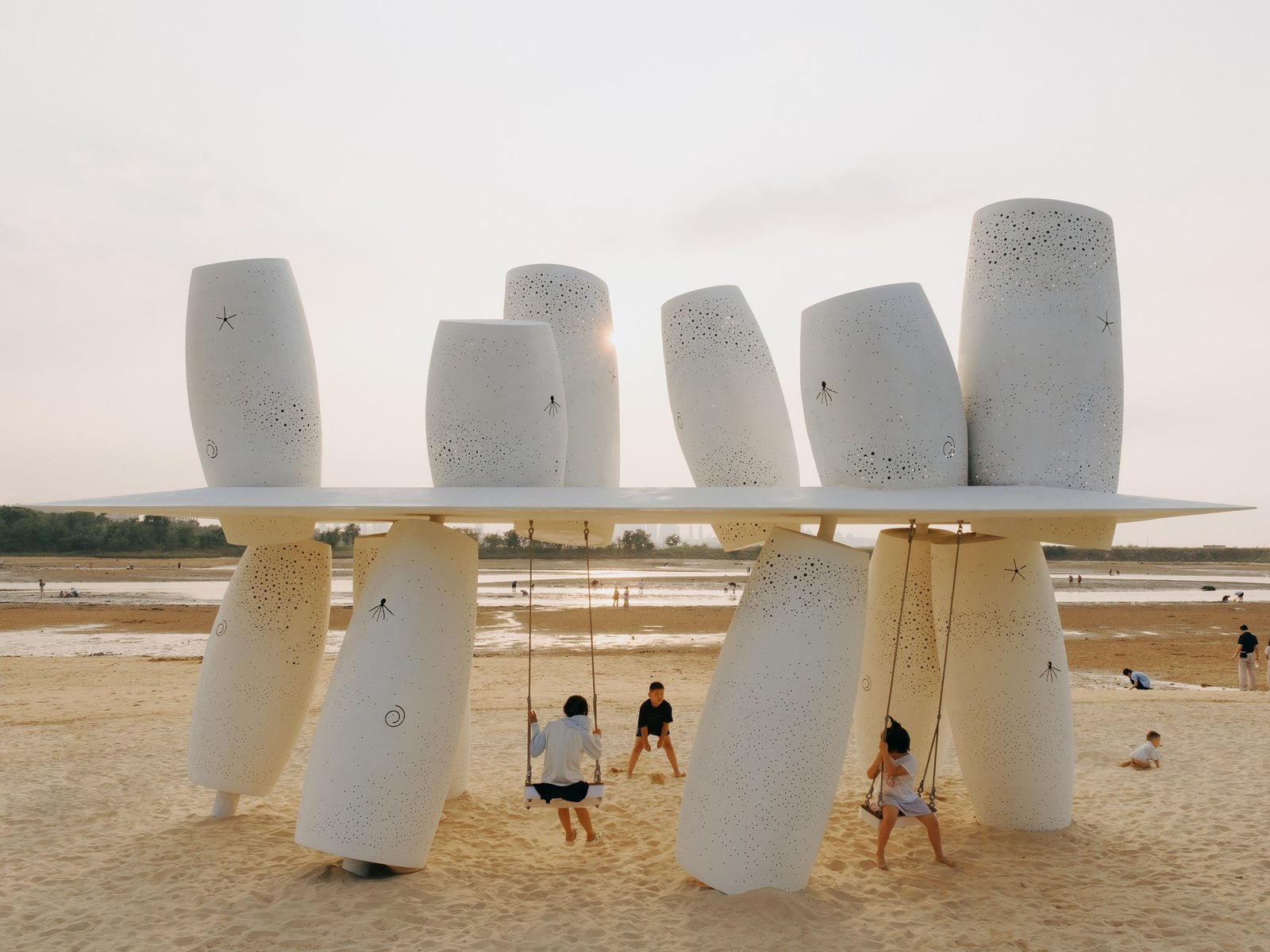 Frontal eye-level view of the Coral Installation featuring perforated white stainless steel cylinders pierced by a thin horizontal roof with children playing on swings on the sandy beach.