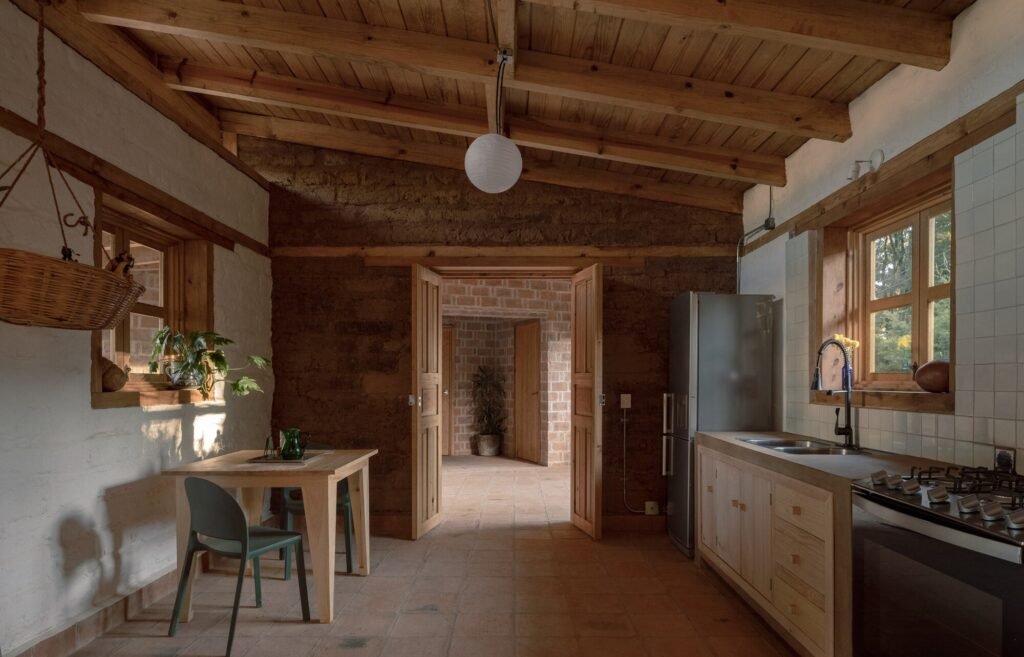 Open-plan kitchen and dining area inside Casa Esteli with exposed wooden roof beams, clay floor tiles, and an open doorway leading to red brick corridors.