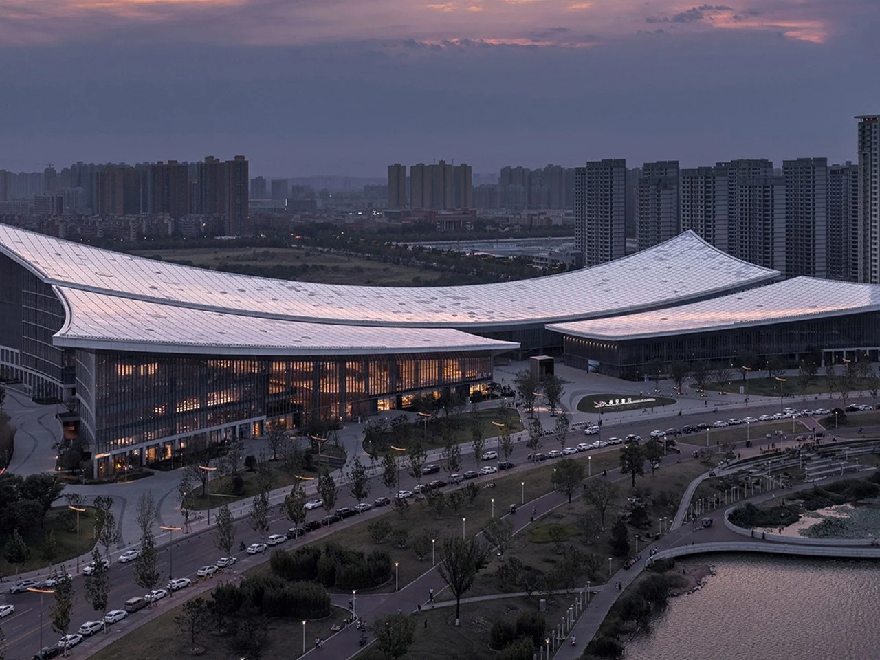 Exterior twilight view of the Xi'an Public Library on the Ba River bank showing its sweeping curved roofline and illuminated glass facade.