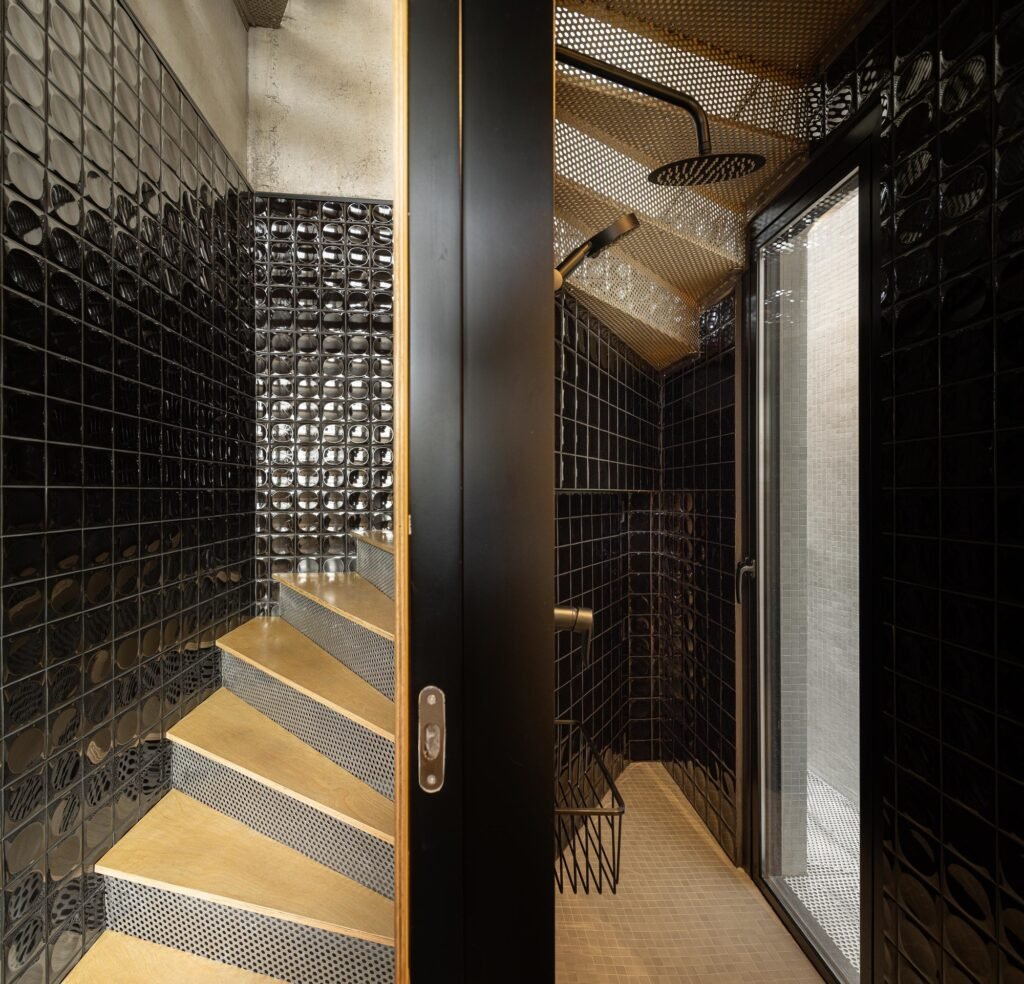 Compact bathroom shower area lined with glossy black square tiles next to a perforated metal staircase and glass opening in Mini Atalhada.