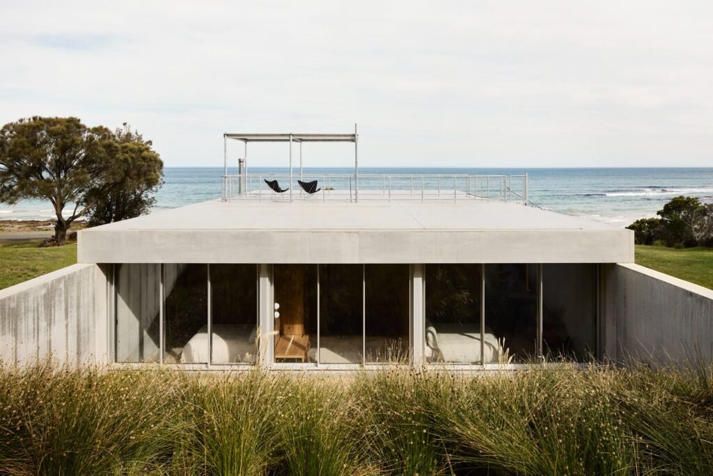 Monolithic concrete structure of Otway Beach House sunk into coastal hill with large glass windows and rooftop deck overlooking ocean.