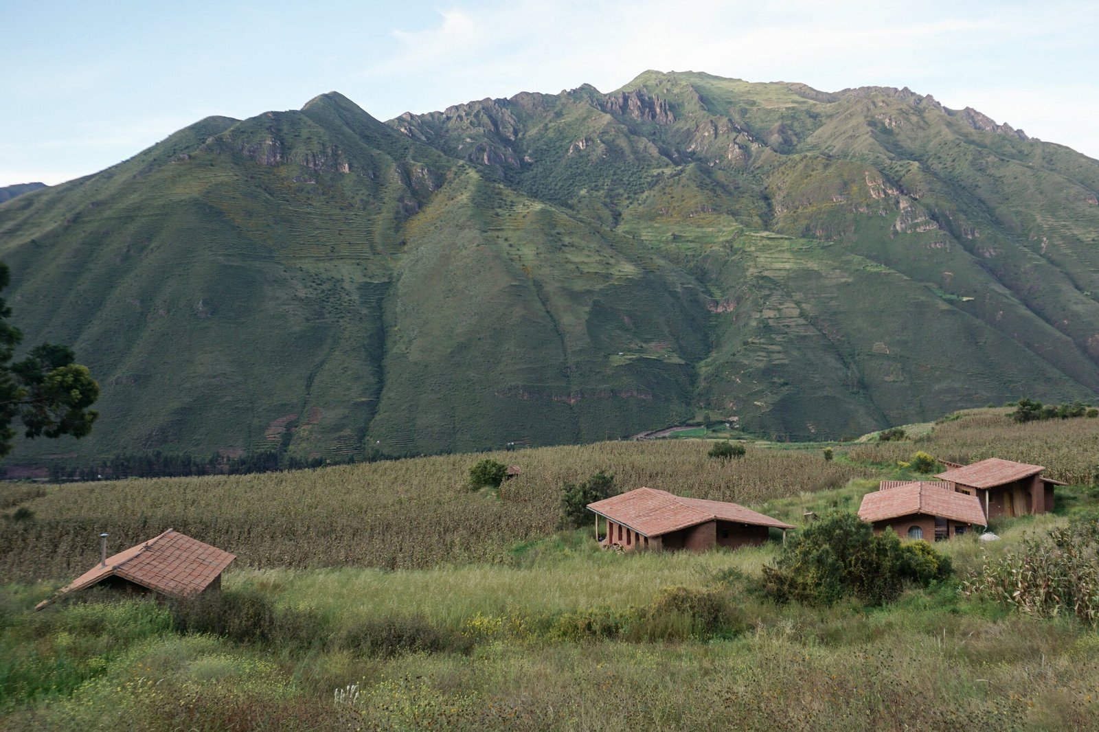 High-angle panoramic view of Kusca project's adobe structures with red tile roofs situated among cornfields against steep Andean mountain peaks near Cusco.