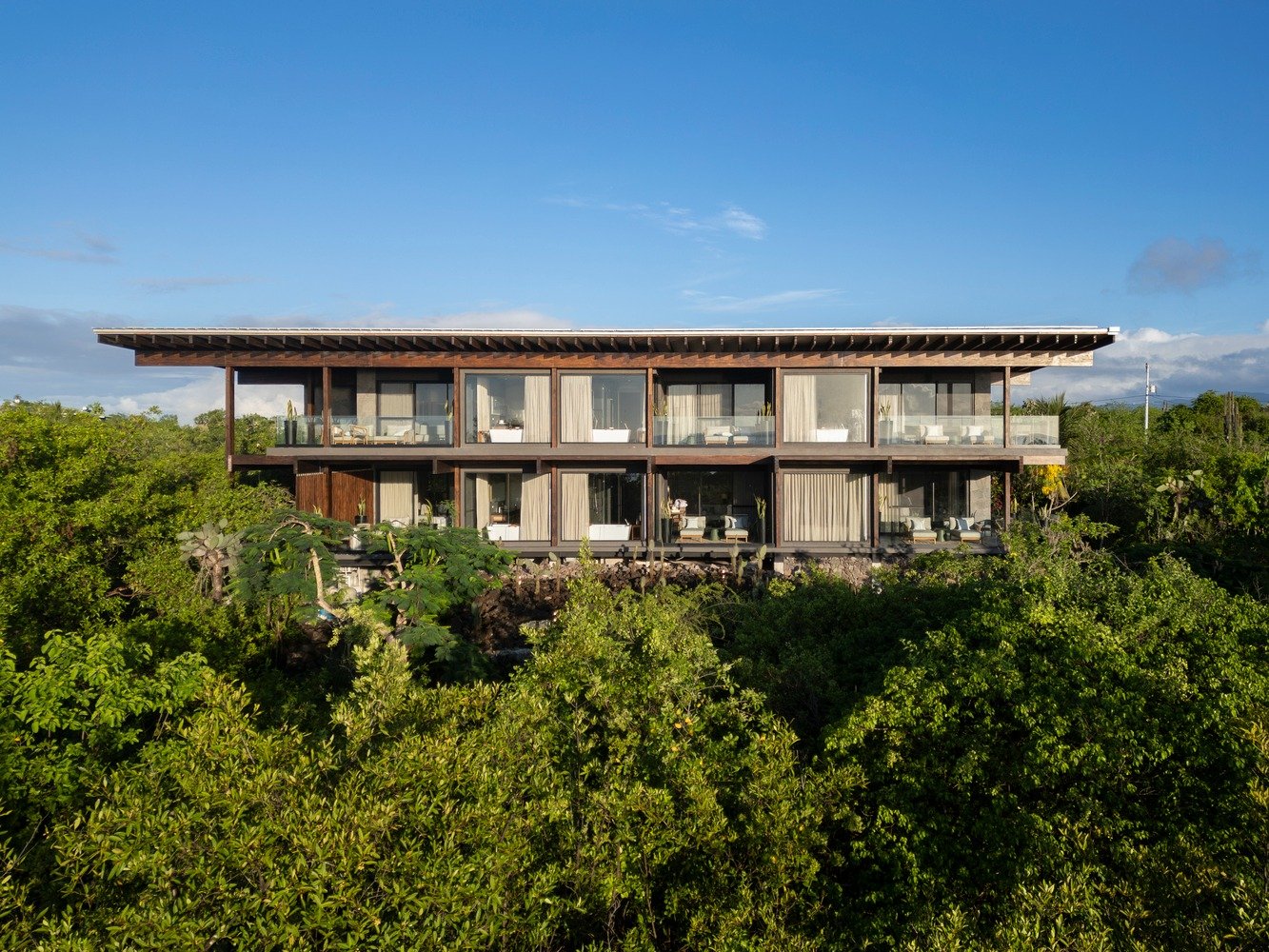 A two-story modern hotel building, constructed with dark wood beams and lava stone base, nestled within dense green mangrove vegetation on Santa Cruz Island. The facade features continuous windows and glass-railed balconies under a blue sky.