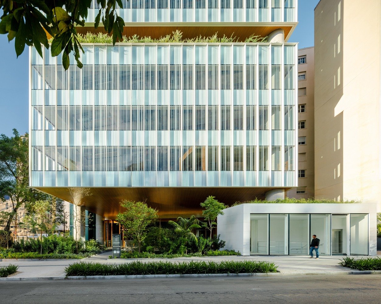 Pedestrian perspective of the Jerônimo Faria Lima building ground level featuring silk-screen printed glass facade panels, warm wooden soffit ceiling, concrete columns, and lush landscaping integrated with the sidewalk.