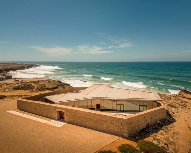 High-angle exterior aerial view of Cresmina Fort in Cascais featuring historic eroded stone outer walls surrounding a modern single-story glass and stone pavilion overlooking the Atlantic Ocean.