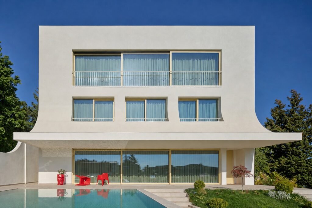 Exterior facade of The Gallery villa featuring pure geometric massing, floor-to-ceiling glass windows, integrated pool, and red terrace furniture.