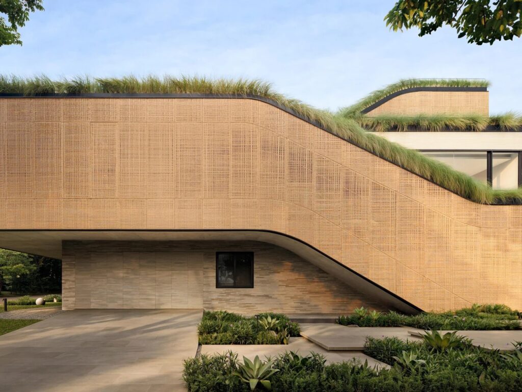 Corner street view of Split Mass house showing woven PE rattan facade screens and a sweeping sloped roofline above the ground entrance.