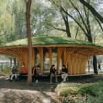 Mushroom-shaped timber pavilion with a green roof surrounded by trees and park visitors resting on an integrated wooden deck bench.