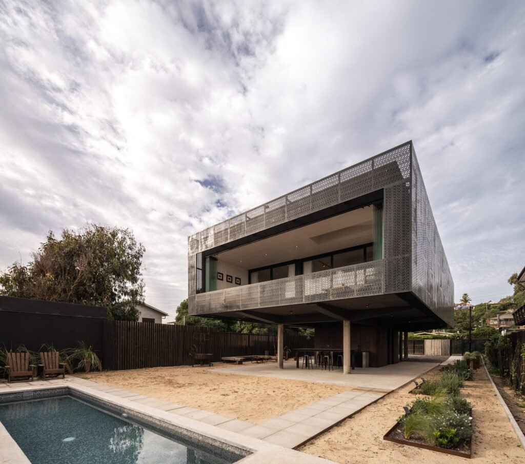Perspective view of Gandarillas House showing the swimming pool, sandy ground level, concrete support columns, and micro-perforated steel facade.