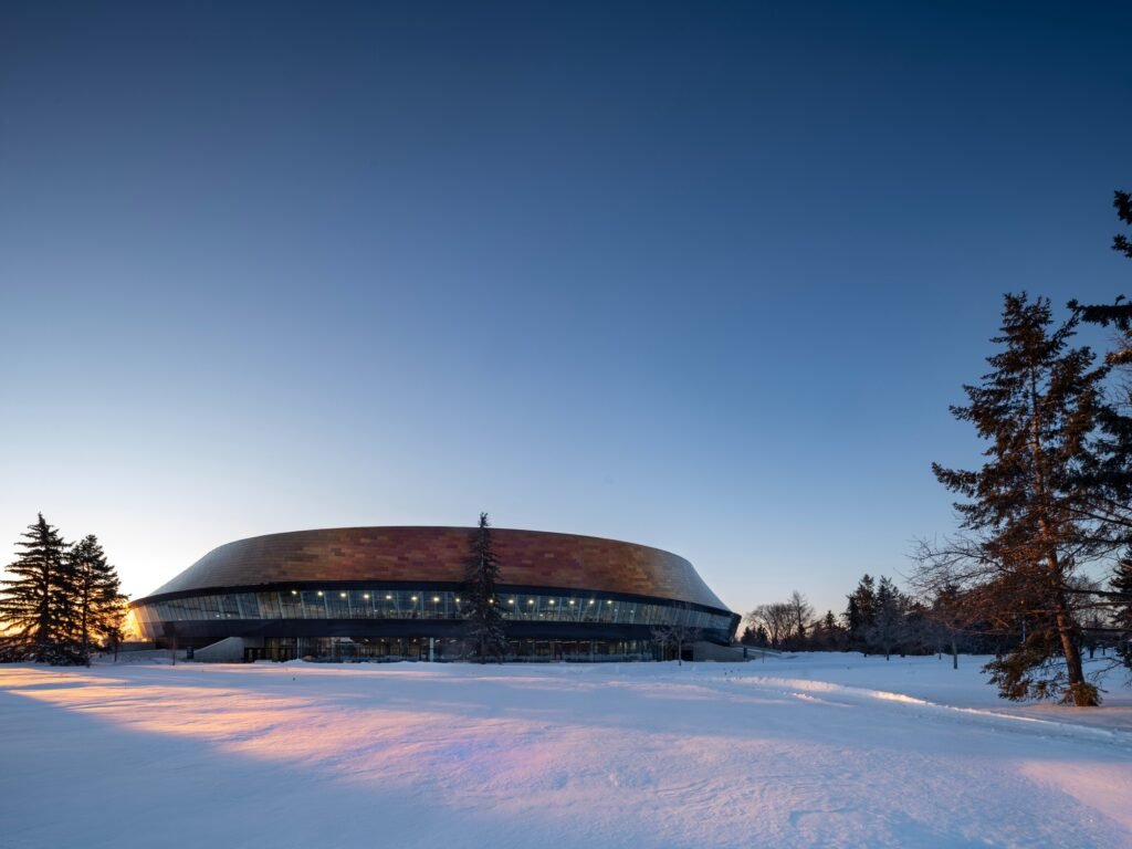 Winter exterior shot of Coronation Park Sports Centre at sunset with snow-covered ground and surrounding pine trees.