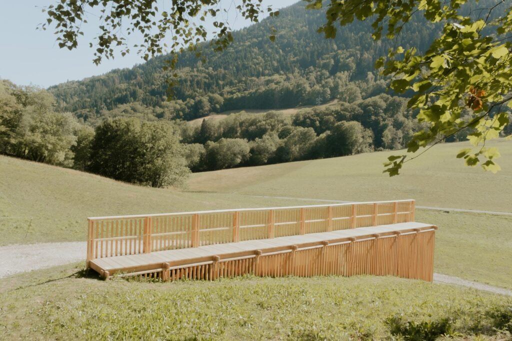 Elevated wooden walkway ramp set against a vast green alpine valley and forested mountain hillsides.