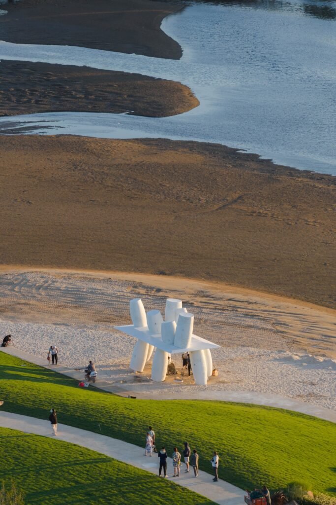 High-angle aerial perspective showing the white Coral pavilion installed on a sandy shoreline adjacent to tidal mudflats, grassy slopes, and a pedestrian pathway.