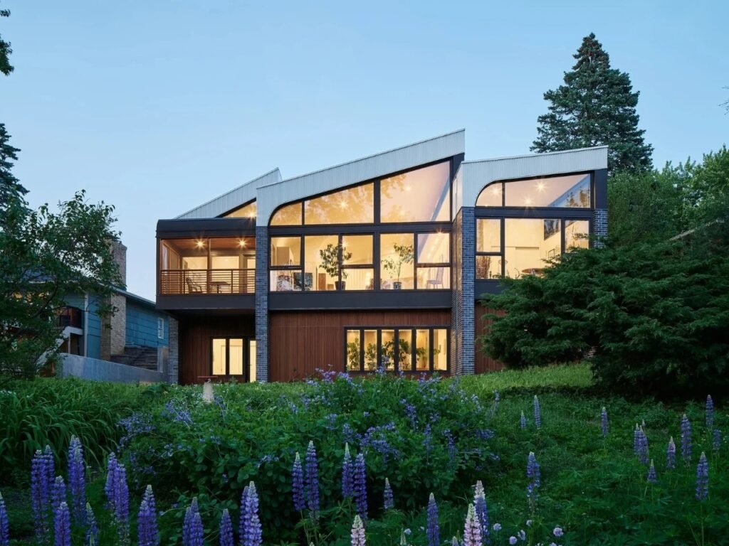 Rear facade of Crescendo house by CityDeskStudio featuring large custom trapezoidal clerestory windows facing a creek and natural landscape at dusk.