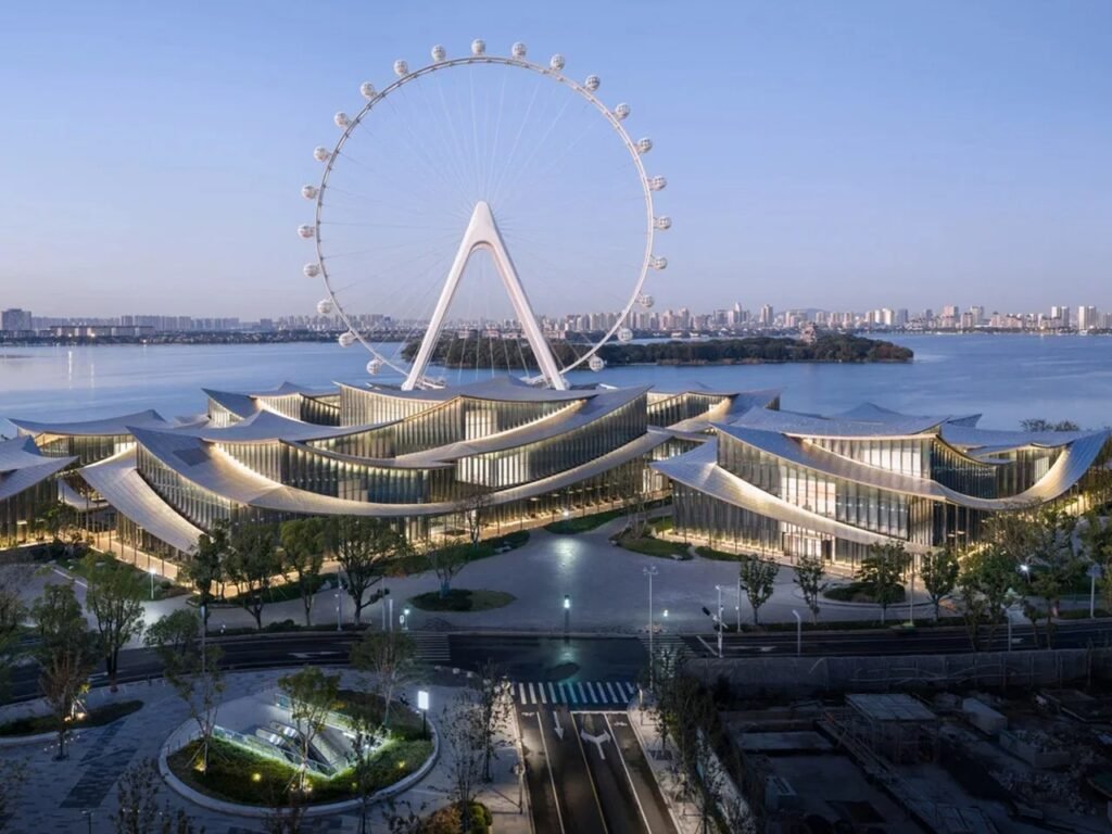 Front ground-level view of Suzhou Museum of Contemporary Art with ripple-like roofs framing a large Ferris wheel behind Jinji Lake.