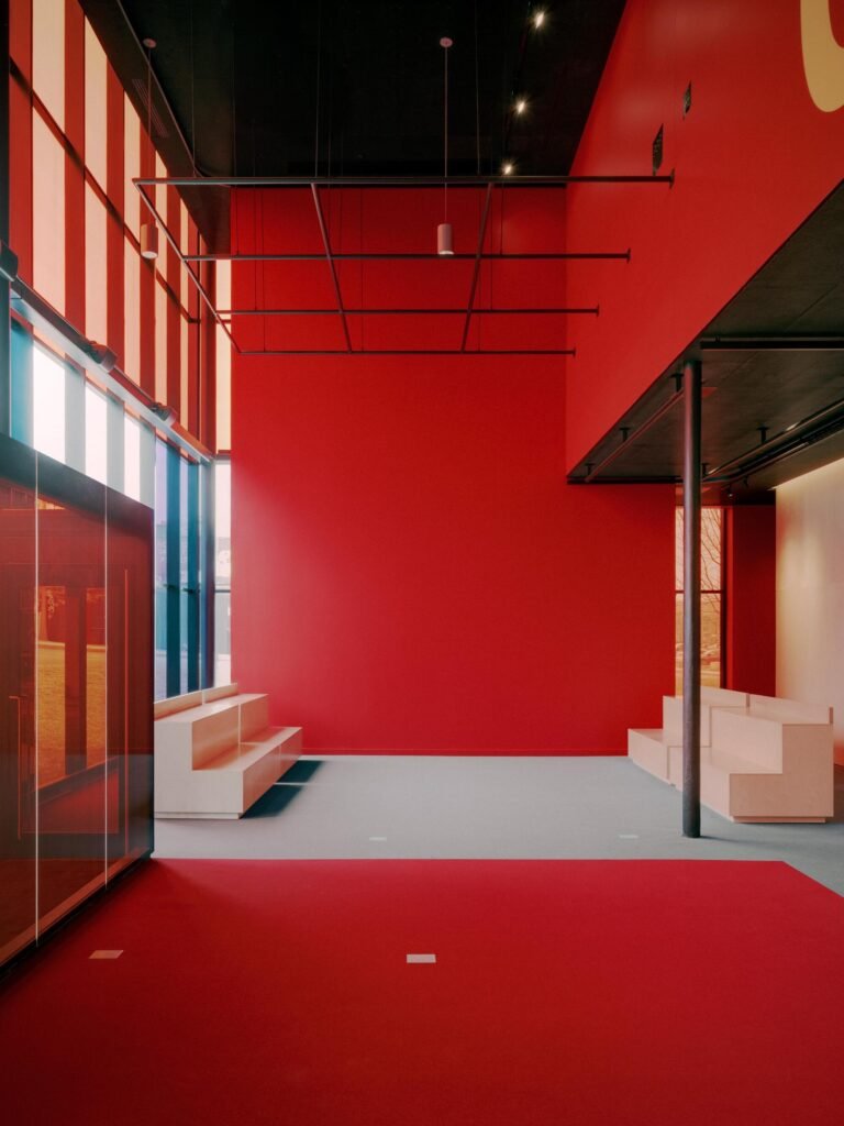 Minimalist double-height interior space in Le Grand-Espace featuring vibrant red walls, light wood modular seating, and overhead technical grids.