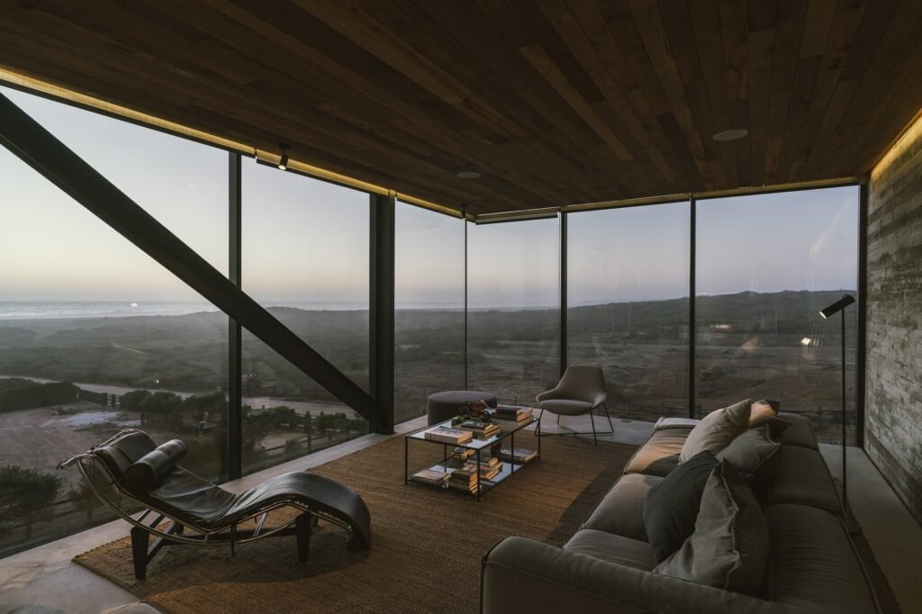 Modern coastal living room inside Casa 22 with floor-to-ceiling corner glass walls offering panoramic ocean views at dusk.