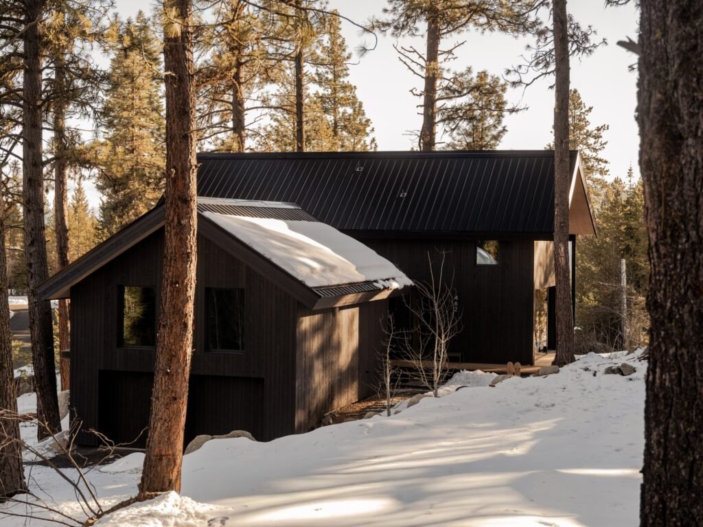 Exterior view of dark gabled cabin nestled in a snowy forest with sloped metal roofs.