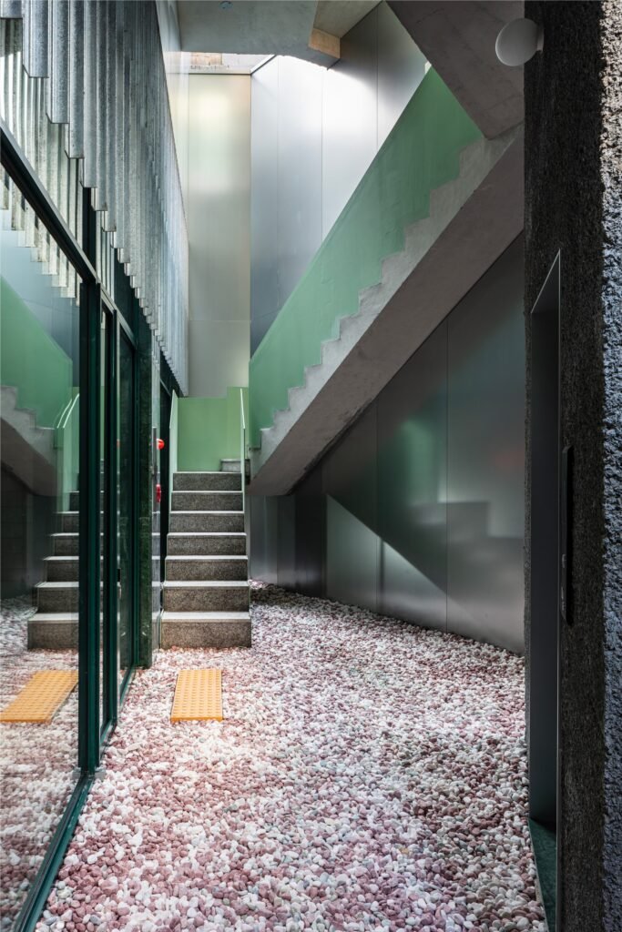 Shared circulation hallway with gravel floor, staircase with green accents, and smooth metal wall panels.