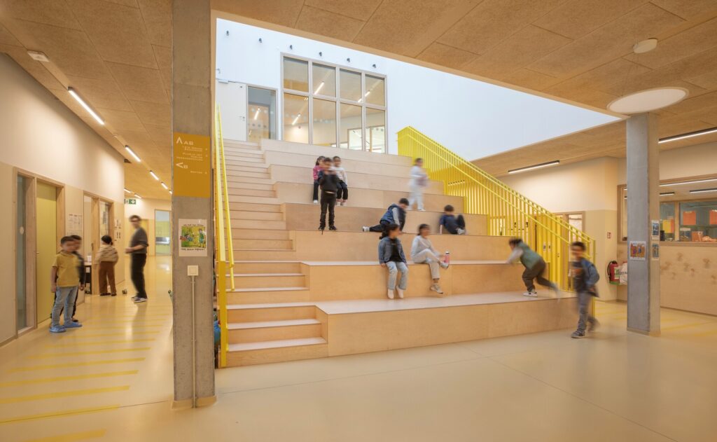Interior central wooden staircase with integrated seating tiers inside Campus Deleers primary school.