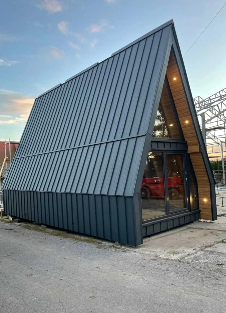Side profile of a contemporary A-frame house showcasing dark grey standing seam metal roof panels and timber-lined front overhang.