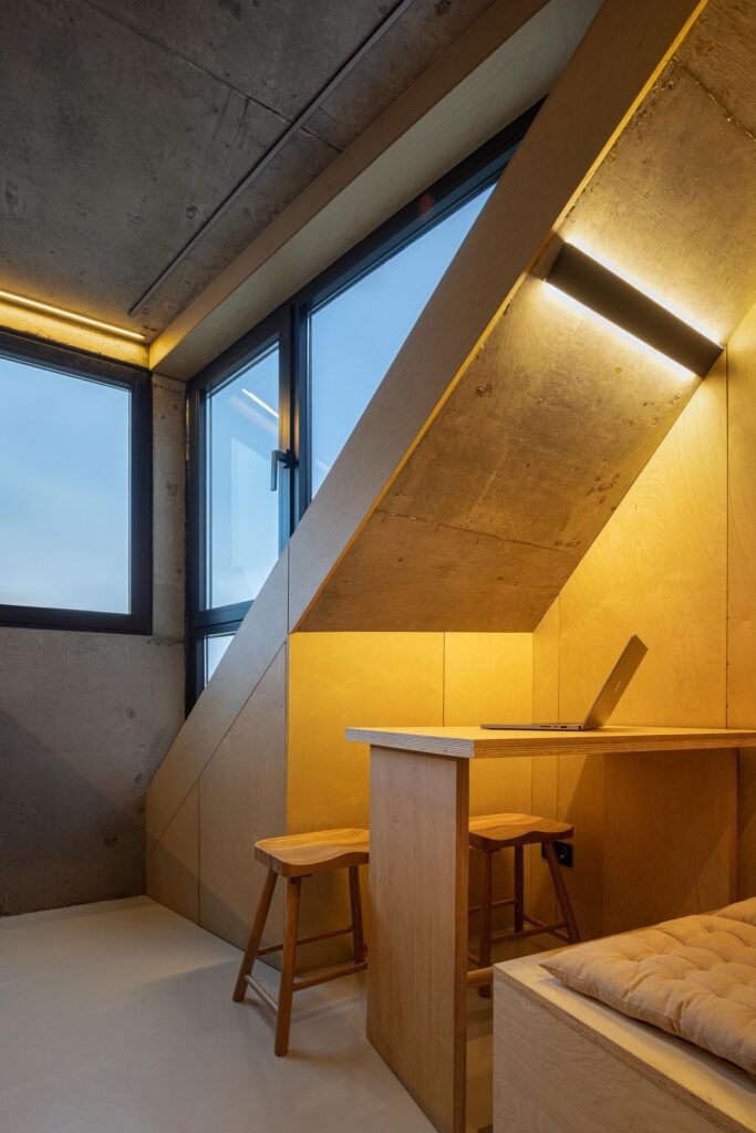 Upper-floor workspace with custom plywood desk, wooden stools, sloped roofline, warm LED strip lighting, and large windows in Mini Atalhada.