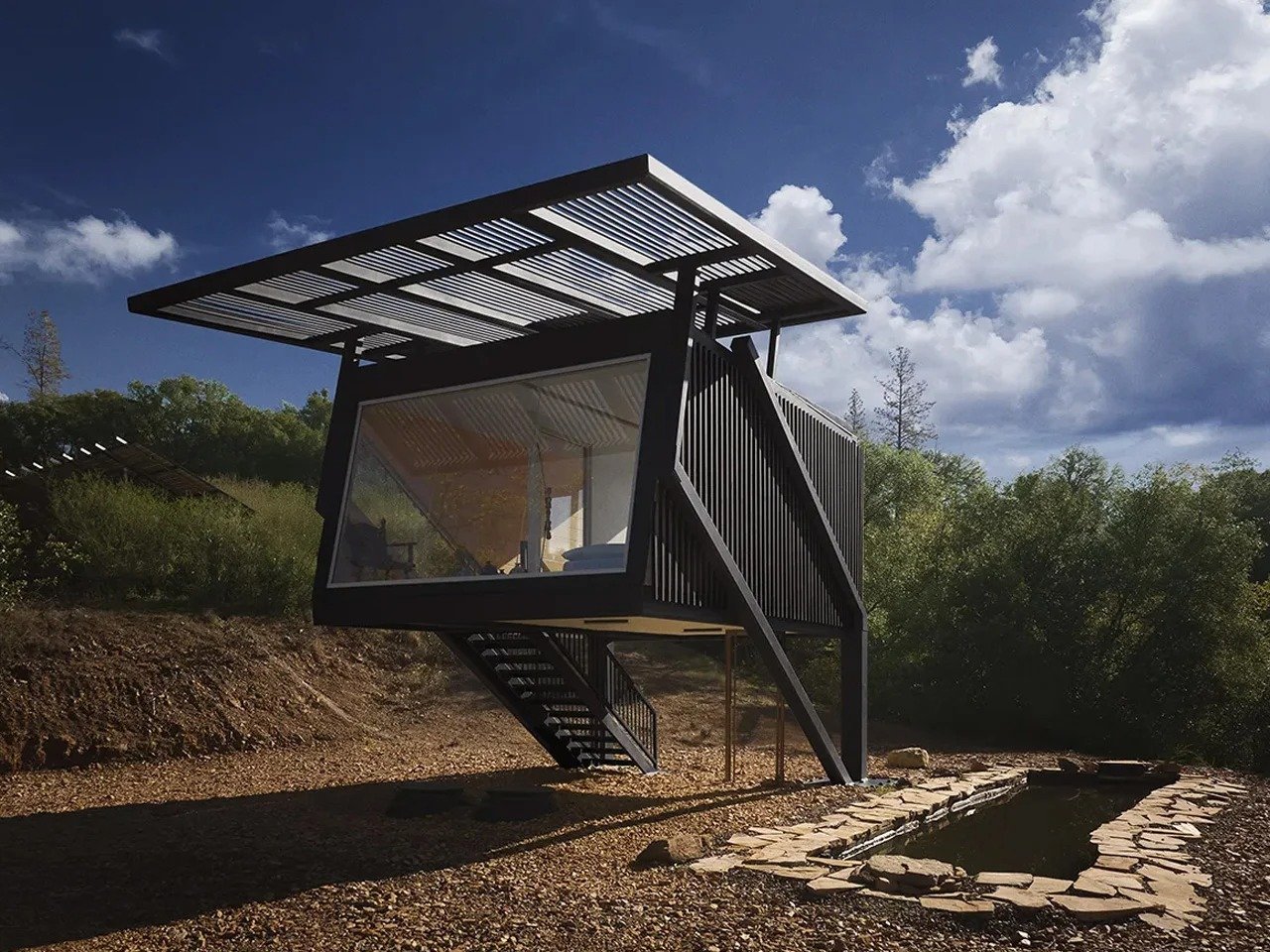 Elevated steel-frame micro dwelling Null House surrounded by trees, featuring a large glass window, external metal staircase, solar canopy, and stone pool.
