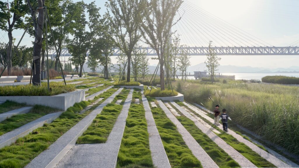Children walking along stepped stone pavers integrated with green grass, leading down toward wild riverfront vegetation and a large bridge.