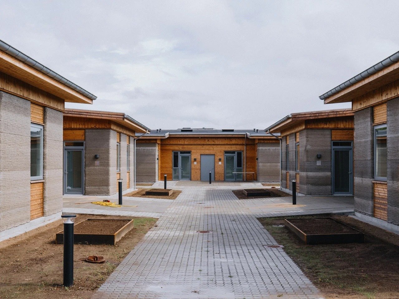 Paved courtyard and central pathway surrounded by low-rise 3D printed concrete student housing units with wood cladding.