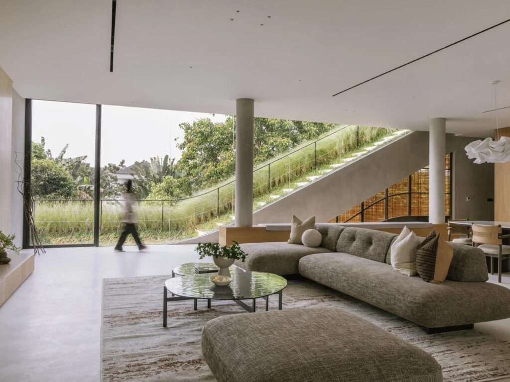 Open-plan interior living room with modern gray sofa looking out through floor-to-ceiling glass to a sloped outdoor grass ramp.