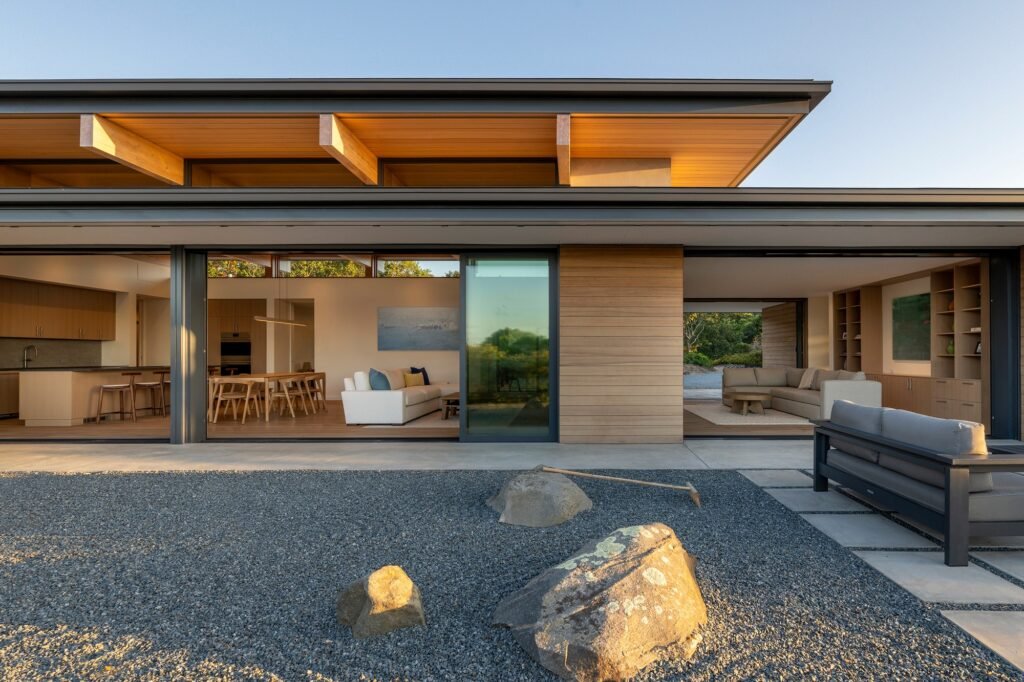 Exterior view of Veil House featuring a gravel Zen courtyard with rocks in the foreground and fully opened glass sliding doors revealing living and dining spaces.