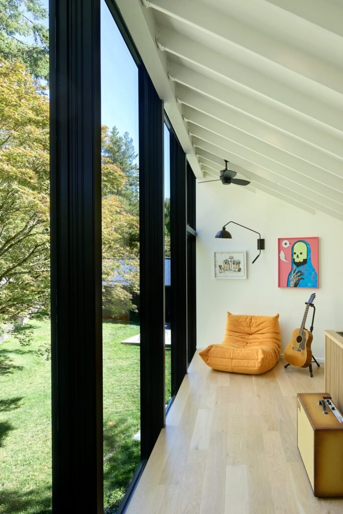 Modern studio interior with sloped exposed ceiling rafters, floor-to-ceiling glass wall overlooking trees, and an orange Togo lounge chair.