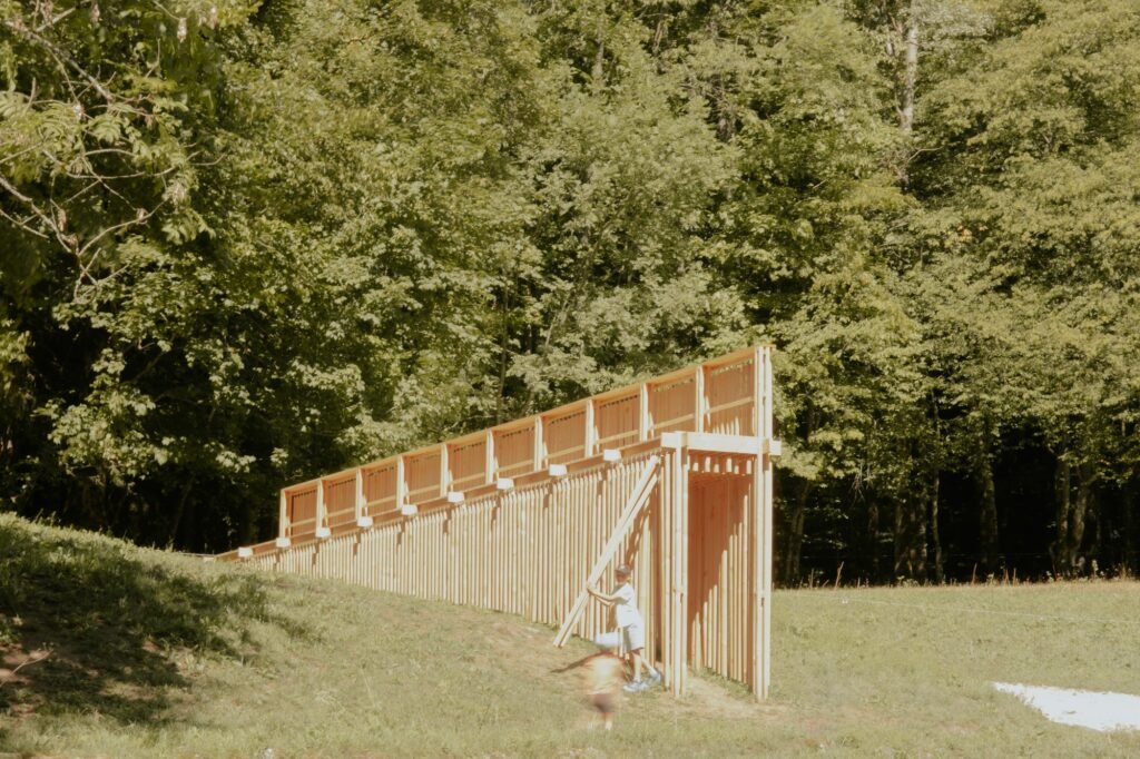 Tapered wooden pavilion structure descending a grassy hill with visitors walking beneath its timber framework near a dense forest.