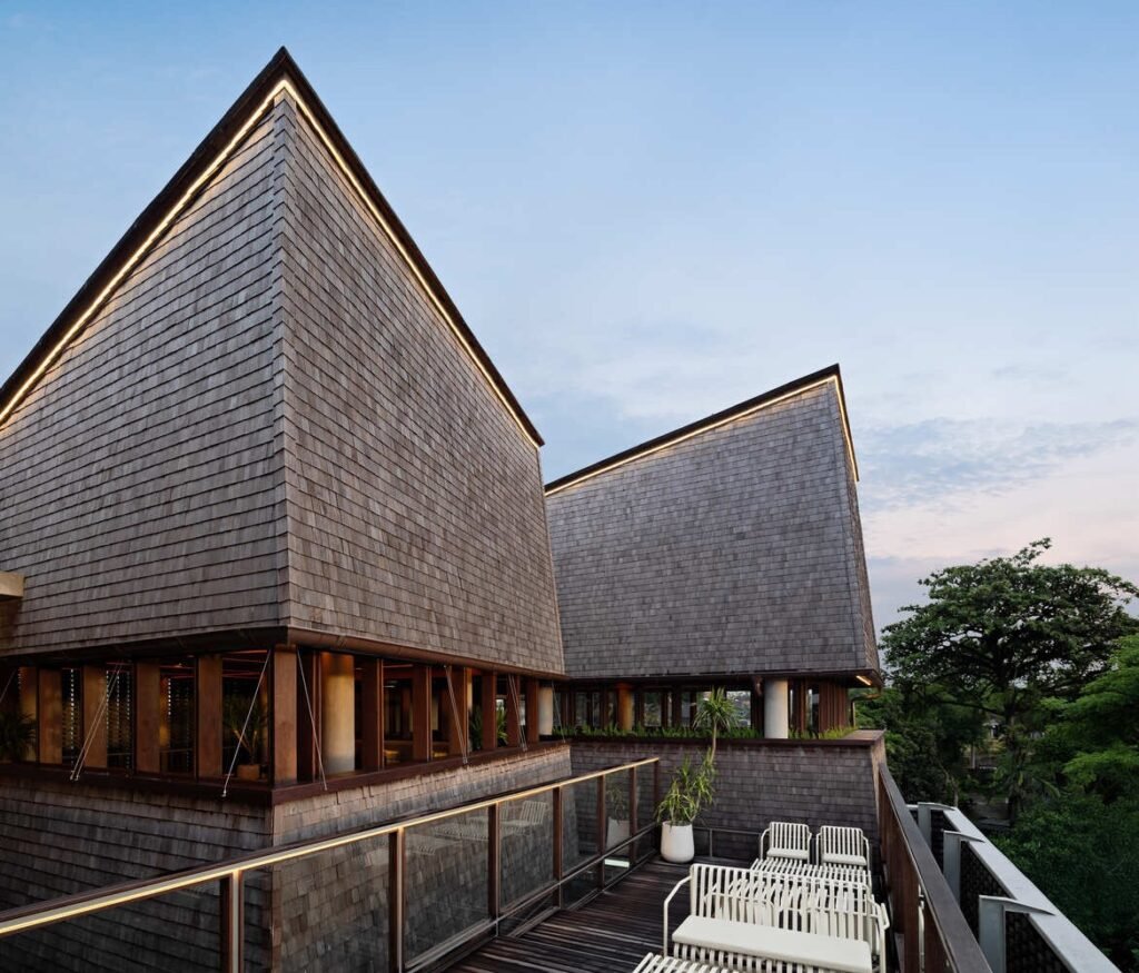 Wooden rooftop deck at Kemilu Ubud featuring contemporary lounge seating under steep shingled roofs against a tropical forest backdrop.