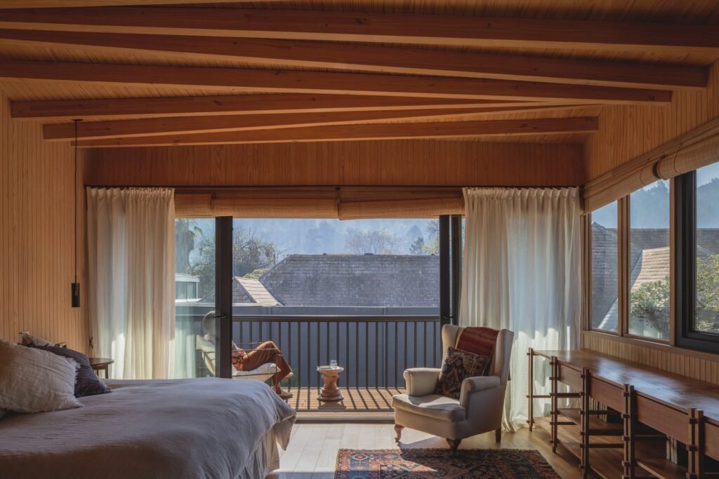 Upper-floor bedroom with wood ceiling beams, large glass windows, and a private balcony overlooking distant hills.
