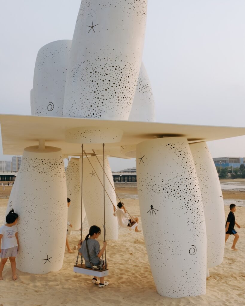 Close-up shot inside the installation showing children interacting with perforated white columns adorned with starfish and sea-life motifs, alongside suspended swings.