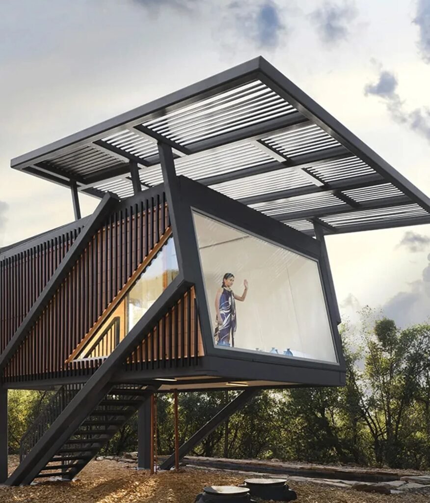 Architect Hafsa Burt standing inside the elevated Null House behind a expansive floor-to-ceiling glass window overlooking woodland.