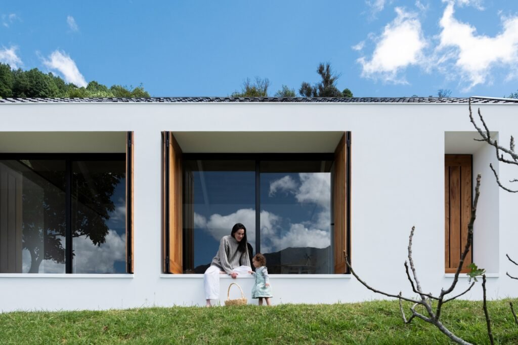 Close-up of the modern facade where a mother and child sit together on a deep recessed window sill flanked by open wooden shutters.