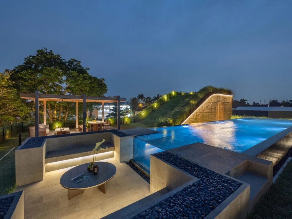 Illuminated rooftop swimming pool at dusk with glass wall edge, sunken seating pit, and wooden pergola lounge.
