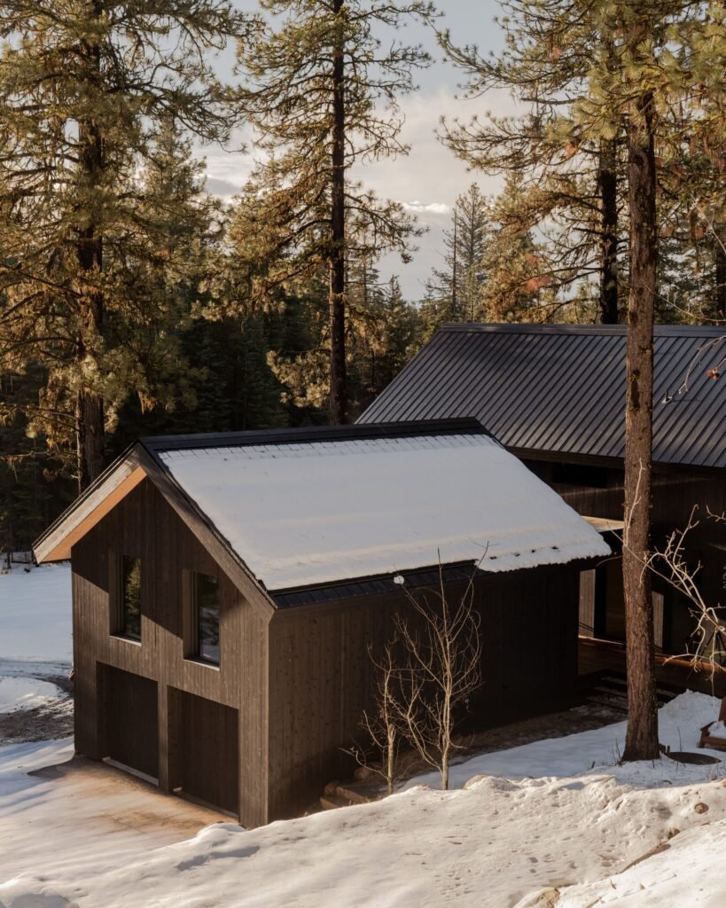 Elevated aerial perspective of a dark gabled cabin garage structure covered in snow surrounded by tall pines.