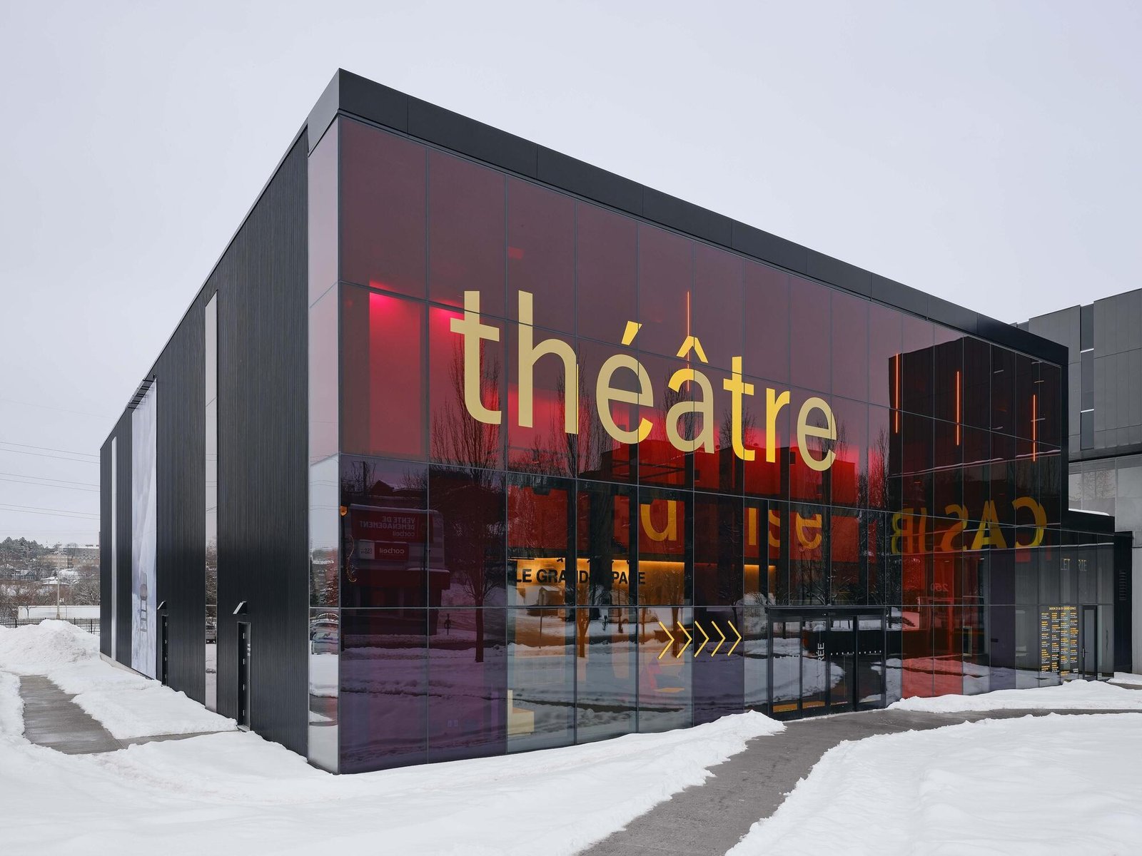 Corner exterior view of Le Grand-Espace displaying prominent 'théâtre' typography on red-tinted glass facade panels in winter.