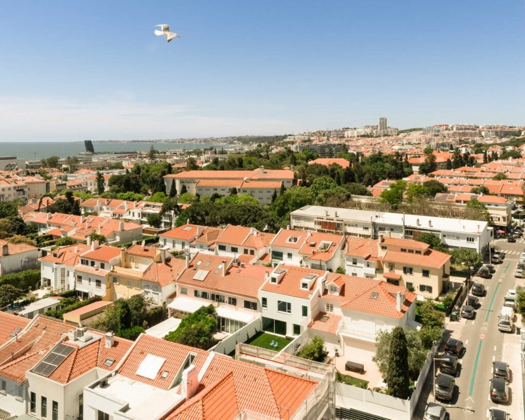 High-angle aerial view of the Restelo housing row in Lisbon, featuring red tile roofs, backyards, and coastal urban scenery.