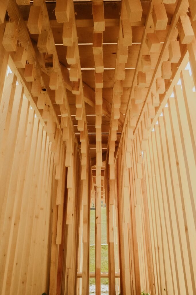 Interior view looking up at dense vertical timber studs and suspended wooden acoustic chimes forming a covered corridor.