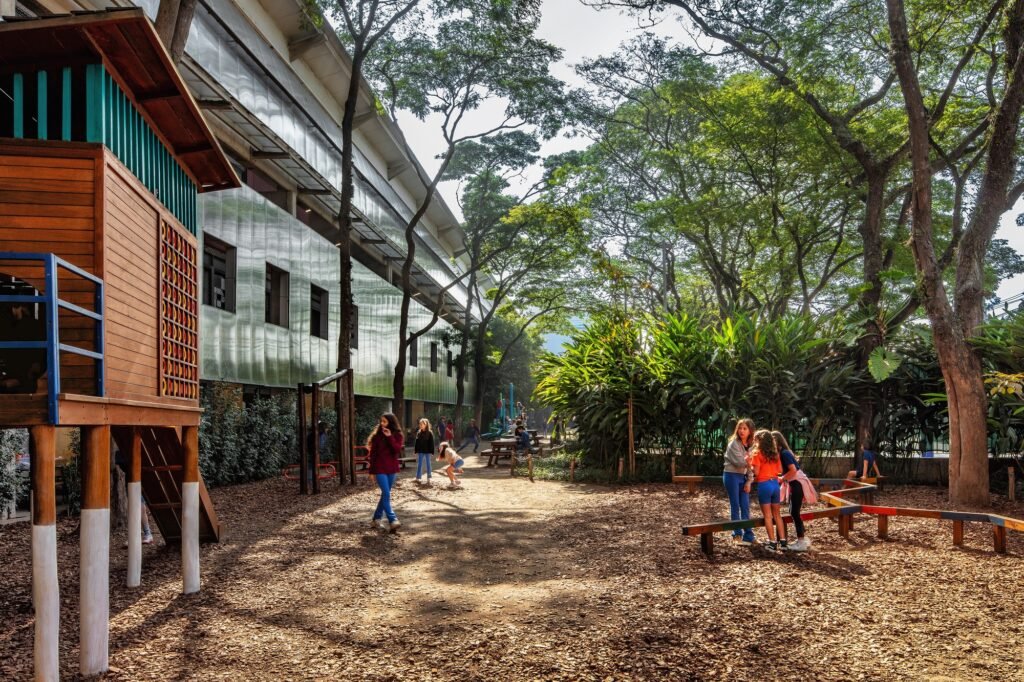Outdoor play area with wooden playground structure, mature trees, and translucent shed facade at Beacon School campus.
