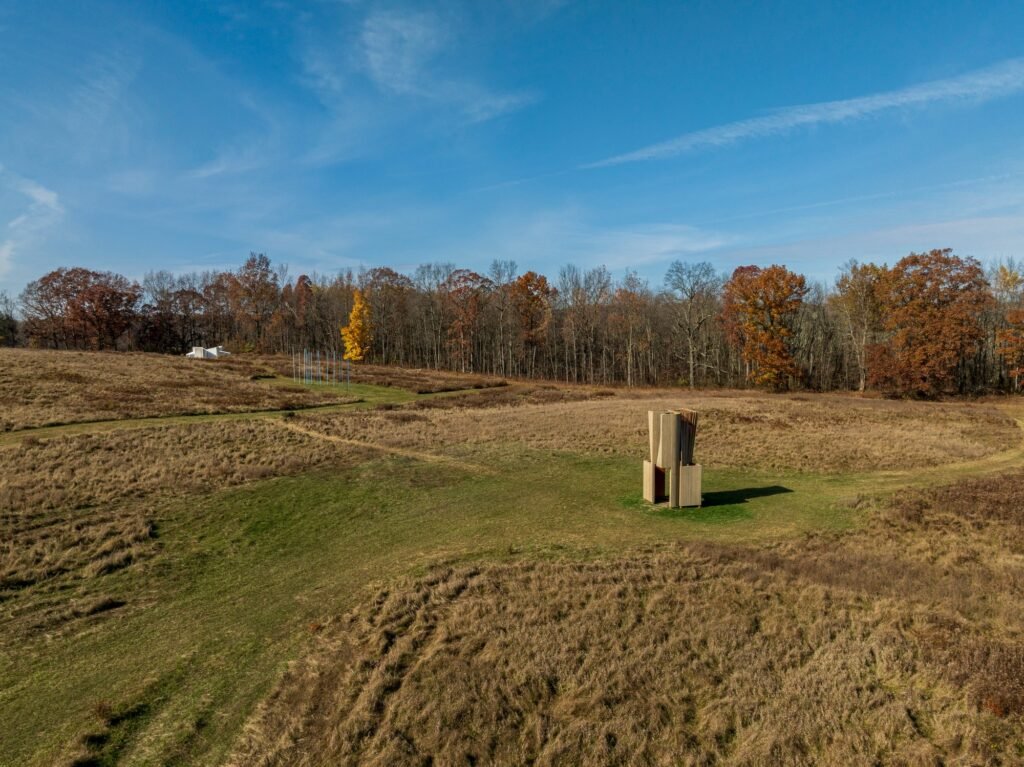 An aerial perspective photo of Match House, a small wooden geometric installation by Young Projects, in a vast, multi-textured field of dry grass with visible dirt tracks, near a line of autumn trees at Art Omi under a blue sky.
